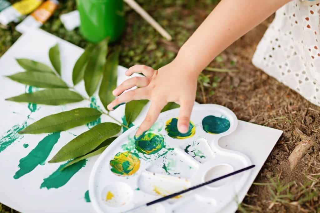 A white piece of paper with a painted leaf print plus a leaf. On top is a paint palette with green paint. A child reaches their hand into the paint to explore making art with plants.